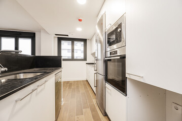 Fototapeta premium kitchen with an island with a black marble countertop, a column of stainless steel appliances, white wooden furniture and wood-like stoneware floors