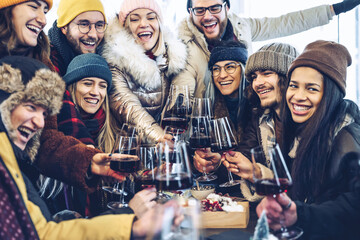 Happy multiracial friends toasting red wine at restaurant pub patio - Group of young people wearing winter clothes having fun at outdoors winebar table - Dining life style and friendship concept