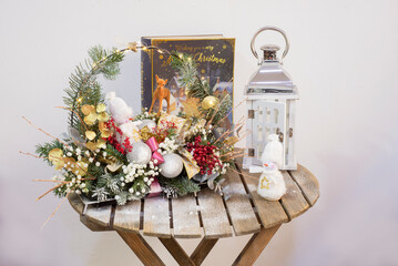 winter New Year's composition for the house from twigs, balls, artificial snow, a luminous garland, a bow, a snowman, a deer a souvenir on a light background on a wooden table with a lamp and a book