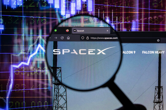 SpaceX company logo on a website with blurry stock market developments in the background, seen on a computer screen through a magnifying glass