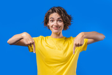 Pretty woman pointing down to advertising area. Blue background. Young lady asking to click to subscribe below. Copy space for your commercial idea, promotional content. - obrazy, fototapety, plakaty
