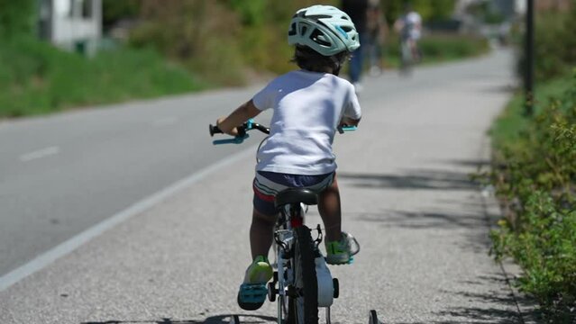 Young boy rides bicycle on green cyclist city road. Back of kid riding bike wearing helmet in summer sunny day