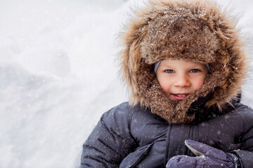 Portrait of a beautiful little Russian boy in a hat with earflaps in winter in the park. Snow is falling. The concept of a happy childhood, Russian winter and lifestyle.