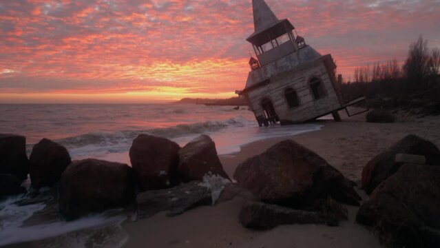 Mysterious vintage flooded leaning wooden house or chapel washed by sea waves, at coastline on pink sunset. Enigmatic rickety building sinking in water on sandy beach near breakwater on sunrise.