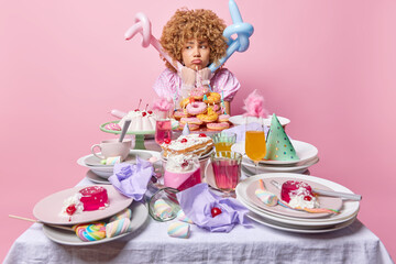 Fototapeta premium Displeased curly haired woman looks unhappily aside feels lonely and sad stands near messy table with delicious desserts holds inflated balloons isolated over pink background. Birthday celebration