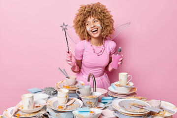 Positive woman dressed in fairy costume does magic for dishwashing poses with wand and bottle of...