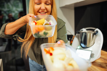 Smiling cute blond mature woman holding, smell fresh vegetables and fruit in plastic containers...