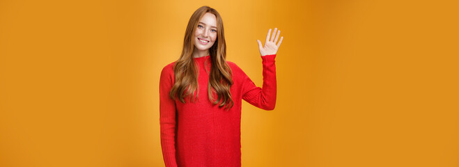 Friendly and optimistic good-looking ginger girl in red sweater raising palm waving at camera,...