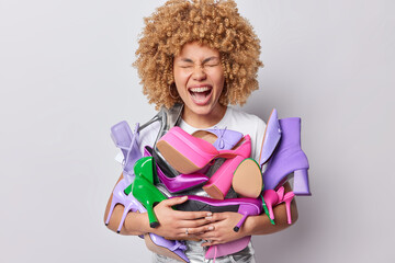 Emotional curly haired woman carries pile of various footwear sorts out her shoes at home being shopaholic and buys fashionable shoes on sale poses indoor against grey background has big collection