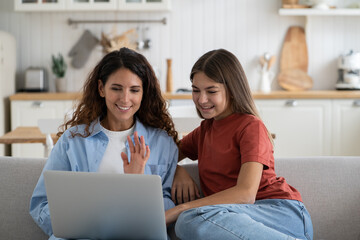 Happy family mother and teen girl daughter waving hand saying hello to relatives while making video call from laptop computer, mom and child sitting on sofa smiling at web camera. Online communication