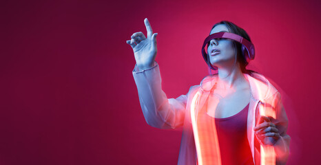 Fototapeta na wymiar Woman in futuristic costume. Girl in glasses of virtual reality while touching air. Augmented reality game, future technology, AI concept. VR. Neon red light. Viva magenta color of the year 2023. 