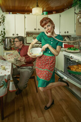 Vintage scene of a wife cooking for a Christmas celebration and talking on the phone, while her husband reads the newspaper sitting at the kitchen table