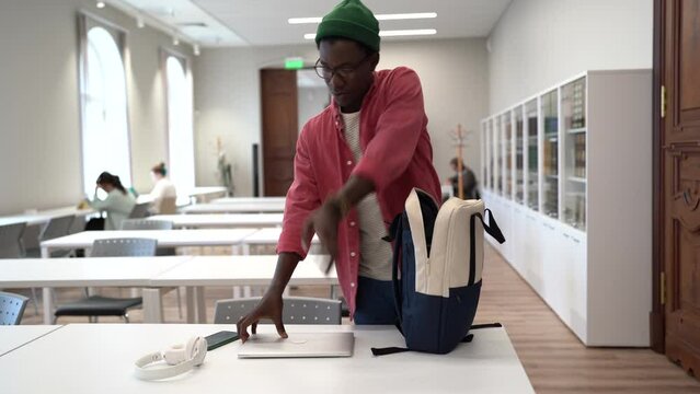 Young African student guy finished study putting laptop and textbook in backpack, packing bag before leaving library. Black man freelancer standing in coworking space finishing workday. Education