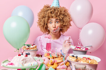Birthday celebration concept. Curly haired woman wears cone hat and dress smeared with cream around mouth drinks cocktail stands near table with desserts comes on party isolated over pink background