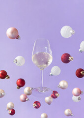 Wine drinking glass with white wine and colorful Christmas baubles against lavender background. Creative party concept. Trendy holiday celebration aesthetic.