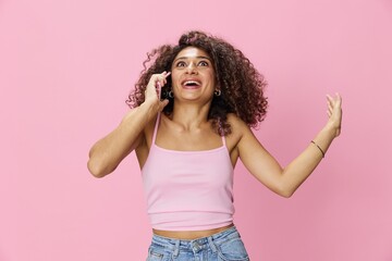 Woman with curly afro hair talking on the phone in a pink top and jeans on a pink background, smile, happiness, finger pointing copy space