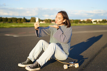 Happy asian girl sits on skateboard, takes selfie with longboard, makes cute faces, sunny day...