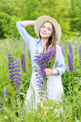 Beautiful young girl in a white dress, straw hat with a bouquet of violet flowers in her hands and picnic basket. Pretty woman in summer in the blooming field holding a bunch of purple lupin