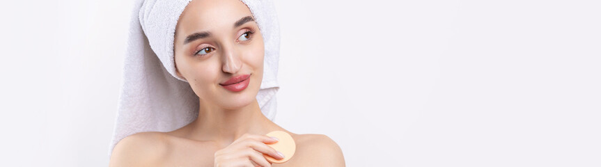 Beautiful girl with thick eyebrows and perfect skin at white background, towel on head, beauty photo. Holds a cosmetic sponge.