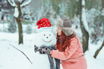 Young pretty girl teenager in fur hat having fun in white winter park among snow-covered trees, going to kiss smiling snowman in christmas red cap and scarf, seasonal outdoor kid's lifestyle portrait