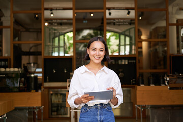 Portrait of asian woman, manager standing with tablet in front of cafe entrance, welcomes guests - obrazy, fototapety, plakaty