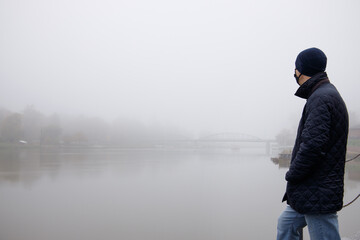  man in mask and hat stands on the bank of the river over which the fog hangs. selective focus 