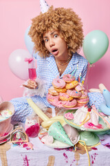 Party and celebration concept. Surprised curly haired young woman wears cone hat and pajama holds glass of cocktail poses near messy table with desserts around with inflated balloons behind.