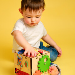 Toddler baby is playing logic educational games busyboard on a studio yellow background. Happy child play with educational toy, learning logic. Kid aged one year four months