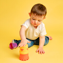 Toddler baby is playing logical educational games with a sandbox mold on a studio yellow background. Happy child playing with an educational toy bucket, learning logic. Kid aged one year four months