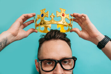 Portrait of an adorable lovely awesome brunette guy holding above his head a golden crown on a blue...