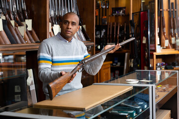 Portrait of hispanic salesman showing collectible old rifled musket on background with rack full of vintage and modern firearms in gun store