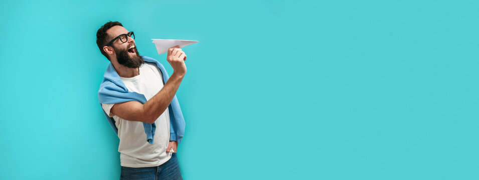Young handsome hipster man with a beard holds a paper airplane in his hand, smiling happily.