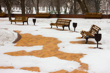 benches of yellow-orange color in a snow-covered park with paving slabs of orange color covered with snow with trees on the background in the park in the winter afternoon