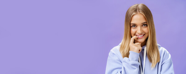 Close-up shot of feminine and tender young girl with fair hair and tanned skin biting finger flirty and shy smiling cute at camera standing in trendy over-sized hoodie, flirting over purple background - obrazy, fototapety, plakaty