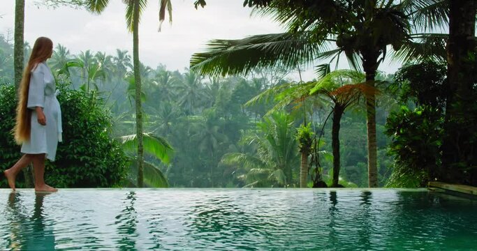 Girl nymph walks along the edge of infinity pool enjoying luxury lifestyle on exotic summer vacation with view of tropical jungle forest in foggy morning.