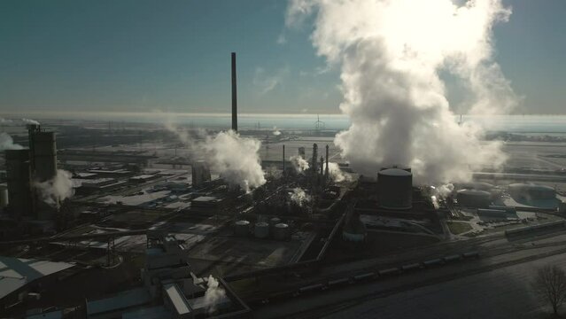 Smoke and steam rising of an industrial facility, CO2 emissions, air pollution, exhaust gases, orbiting aerial drone shot, backlit scene