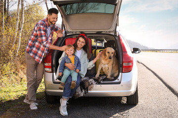 Parents, their daughter and dog sitting in car trunk near road. Family traveling with pet