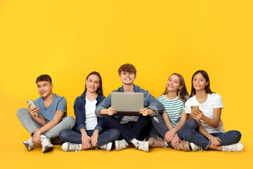 Group of happy teenagers with laptop and smartphones showing thumbs up on orange background
