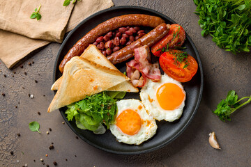 English Breakfast with two eggs, sausage, bacon, toasts and vegetables, red beans on a black plate top view