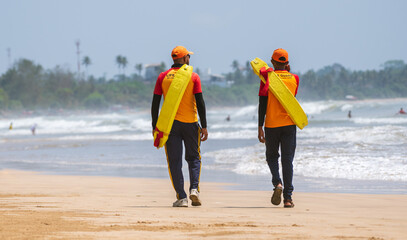 Two Lifeguards on duty at the tropical beach, Both carrying rescue tubes on their shoulders and...