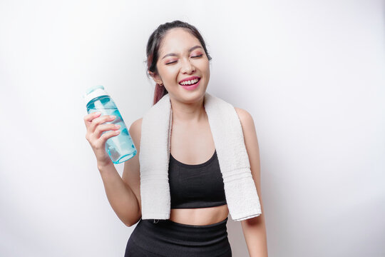 Sportive Asian woman posing with a towel on her shoulder and holding a bottle of water, smiling and relaxing after workout