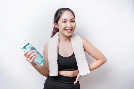 Sportive Asian woman posing with a towel on her shoulder and holding a bottle of water, smiling and relaxing after workout