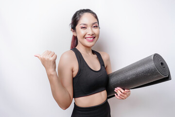 Excited sporty Asian woman wearing sportswear pointing at the copy space beside her while carrying yoga mat, isolated by white background