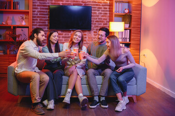Obraz na płótnie Canvas Five friends sitting on a sofa in a cozy room and clink paper cups with beverages