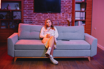 Obraz premium Young woman sitting on a sofa in a cozy room and looking directly at camera