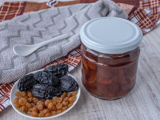 A jar of white cherry jam and a plate of dried fruits, raisins and prunes, a white glass teaspoon. Close-up side view from above. The concept of choosing healthy or unhealthy sugar.