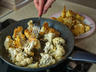Close-up of female hands with a fork laying out pieces of fried cauliflower from a frying pan onto a plate. A housewife puts pieces of cooked fried cauliflower from a frying pan onto a plate
