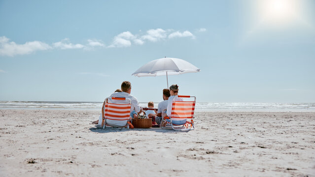 Family, relax and picnic in the sun on the beach for summer vacation, holiday or weekend getaway in the outdoors. People relaxing by the ocean coast with chairs and umbrella for free time in nature