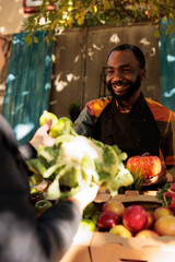 Fototapeta premium Cheerful farmer vendor presenting locally grown food on farmers market stand, small business organic products. Young man talking to senior farming customer, various fresh bio produce.