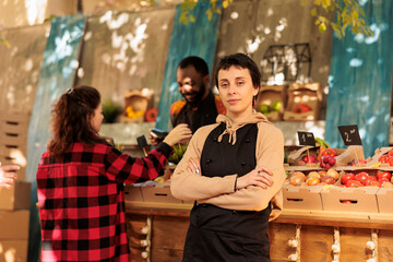 Cheerful young farmer posing next to farmers market stall, presenting healthy food natural products outside. Confident woman selling seasonal organic produce, fruits and veggies.